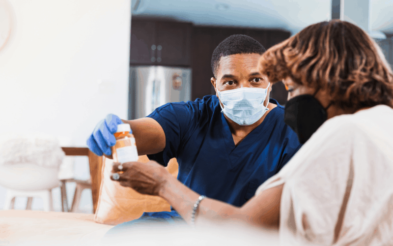 During a home health visit, the Black male nurse hands the senior woman her medication. They are both wearing masks to prevent the transmission of the coronavirus.